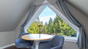Una habitación con techo inclinado, una mesa redonda con dos sillas, un florero con flores y una ventana con cortinas.