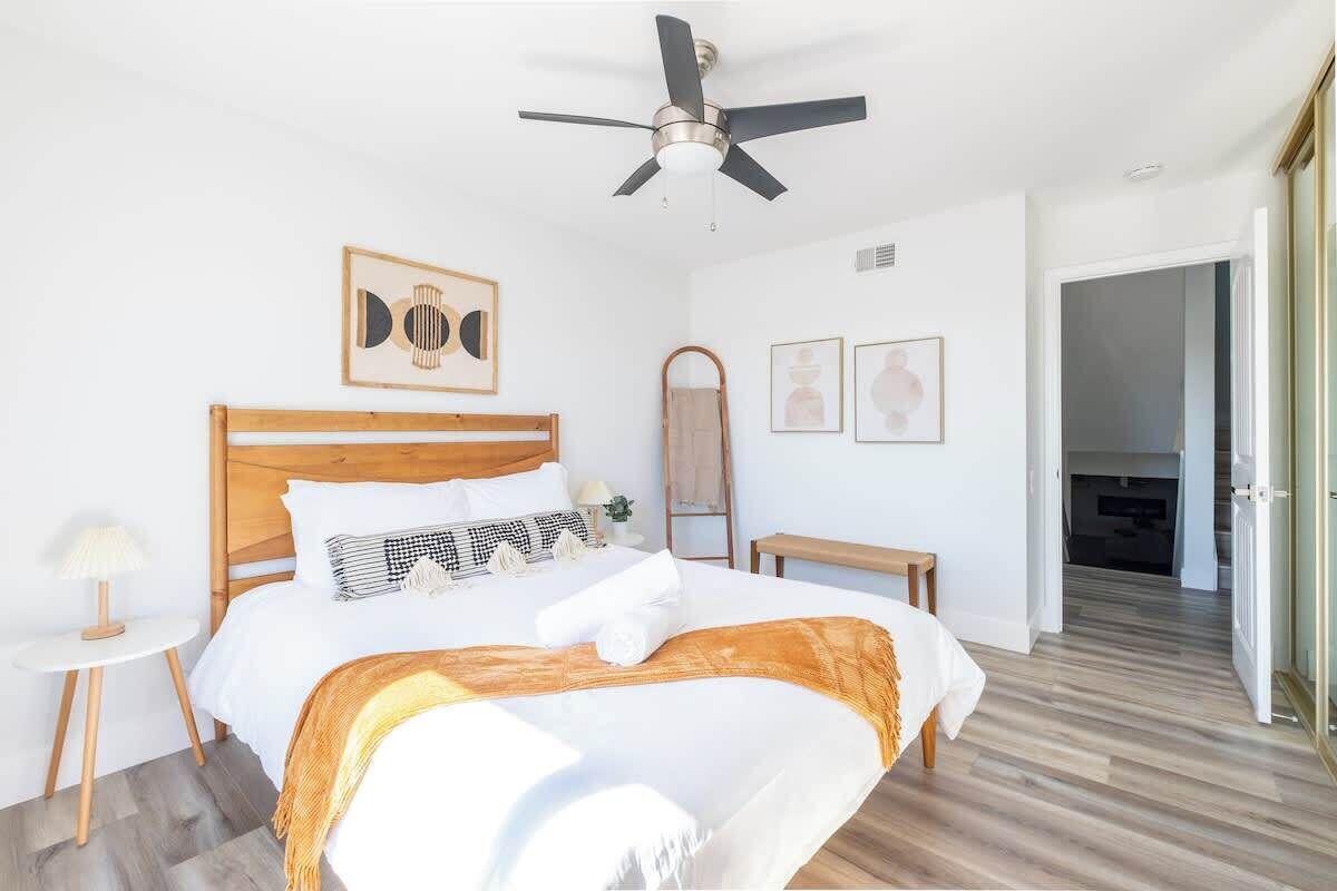 Un dormitorio con cama de madera, ventilador de techo, mesita de noche con lámpara y un espejo en la pared.