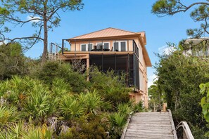 A modern house with a wooden deck leading to a glass entrance, surrounded by lush greenery.