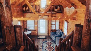 A wooden cabin interior with a staircase leading to a loft area, a dining table, and a blue patterned rug. - Charming Lakeview Cabin Near Geneva-On-The-Lake, The Perfect Getaway! (Ashtabula)