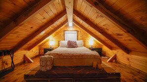 A cozy attic bedroom with a sloped wooden ceiling, a large bed with a patterned blanket, two bedside lamps, and a window with blinds. - Charming Lakeview Cabin Near Geneva-On-The-Lake, The Perfect Getaway! (Ashtabula)
