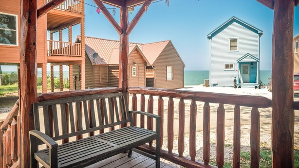 A wooden bench on a covered porch overlooking a beach with a white building and smaller structures. - Charming Lakeview Cabin Near Geneva-On-The-Lake, The Perfect Getaway! (Ashtabula)