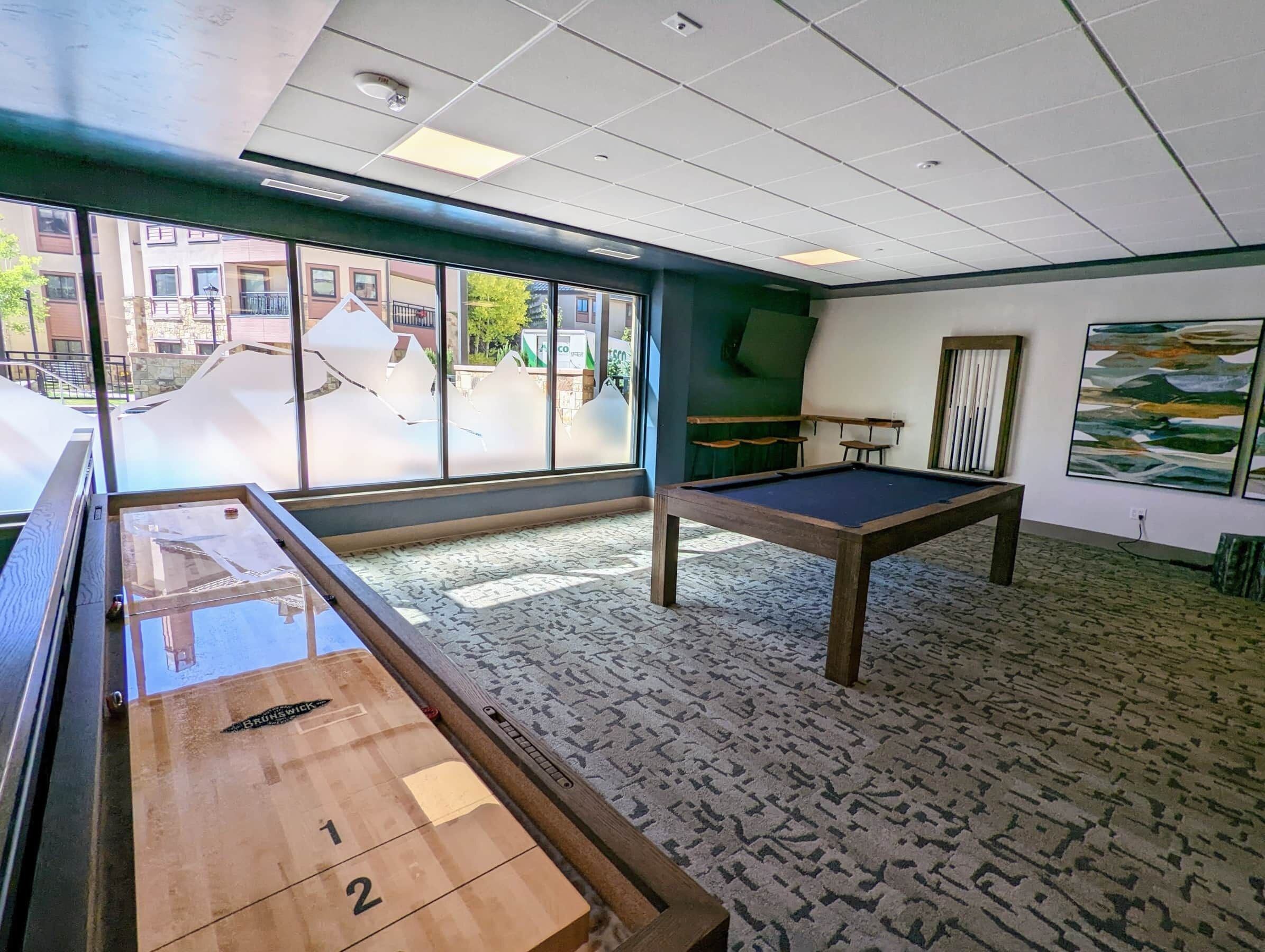 A pool table, shuffleboard table, and artwork in a room with large windows and patterned carpet.
