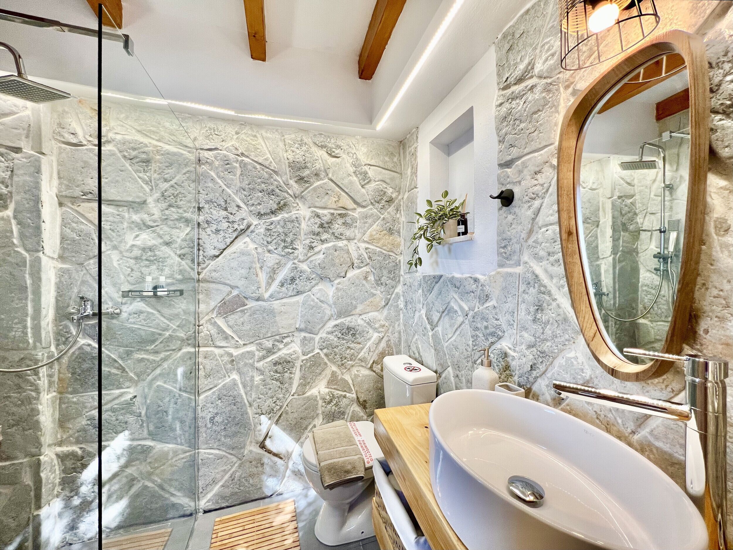 Un baño con pared de piedra, cabina de ducha de vidrio, tocador de madera y un espejo grande.