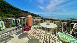 A balcony with a table and chairs, a potted plant, and a view of a mountainous landscape and sea.
