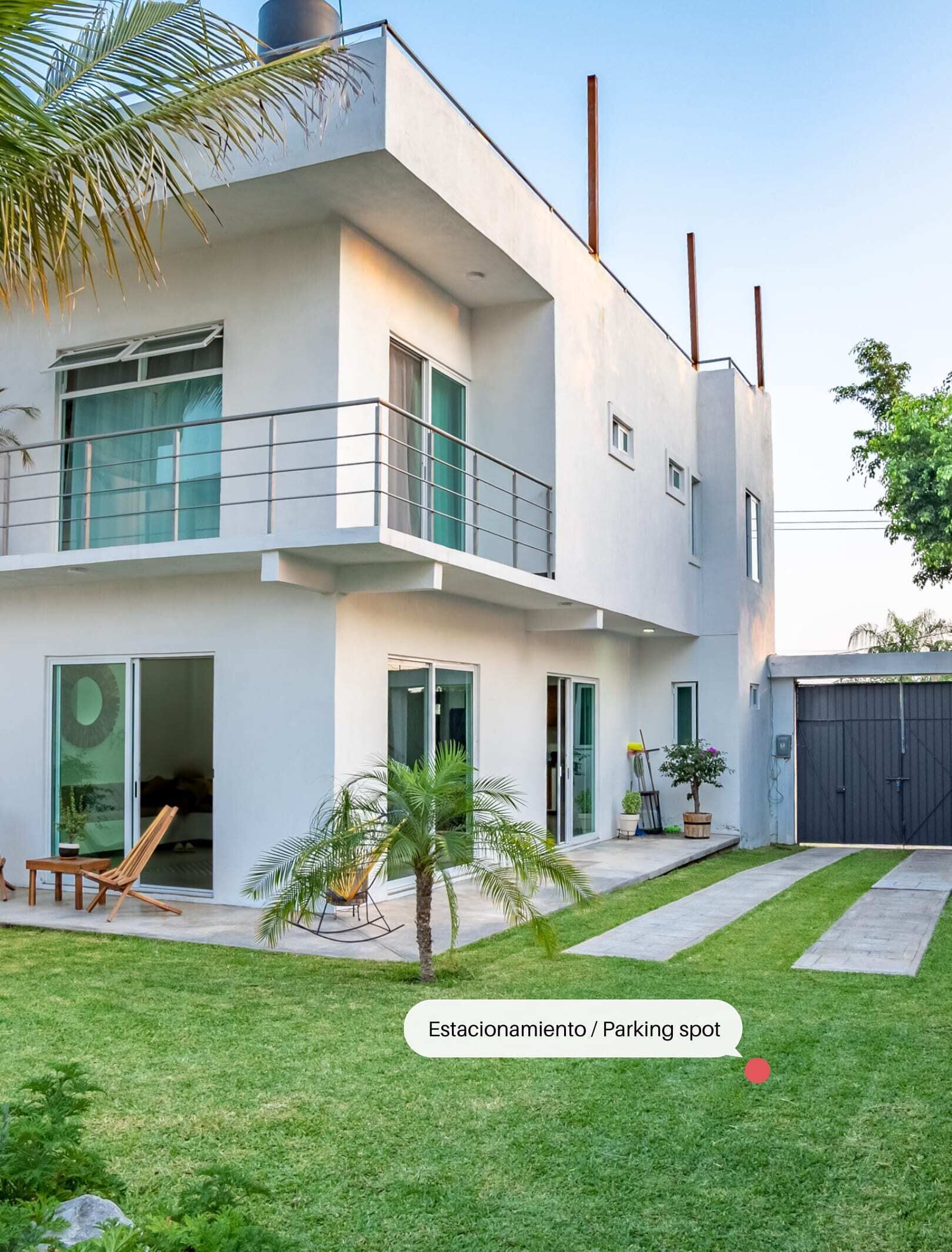 Une maison moderne à deux étages, dotée d’un balcon, de grandes fenêtres et d’un petit jardin.