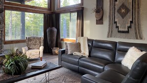 Een ruime woonkamer met een leren bank, een bijzettafeltje en een groot raam met uitzicht op het bos.