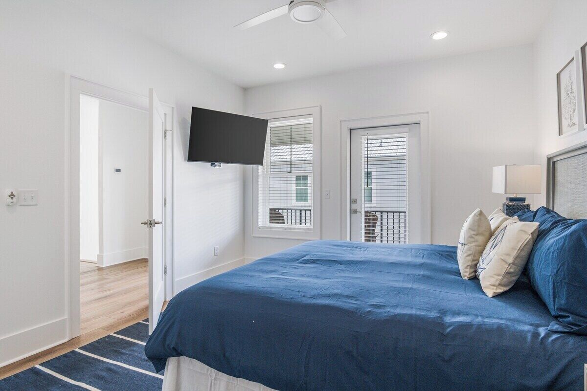 A bedroom with a large bed, a ceiling fan, a flat-screen TV, and a balcony with blinds.