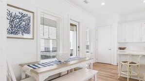 A white interior with a wooden dining table, white chairs, and a bench. There is a framed artwork on the wall, a kitchen area with white cabinets, and a window with blinds. - Lost Dunes Cottage- Walking Distance to Beach! (Gulf Shores)