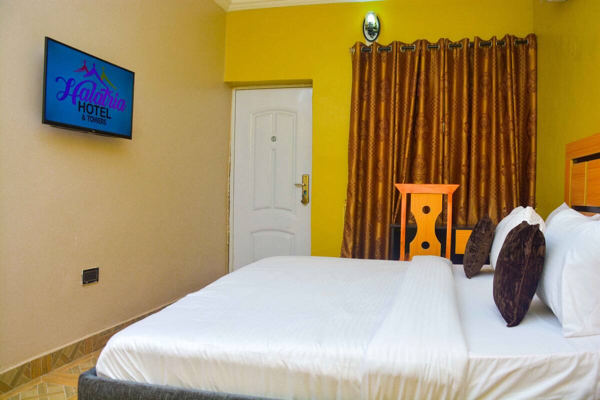 A hotel room with a bed, a television displaying the hotel name, a door, and a curtain.