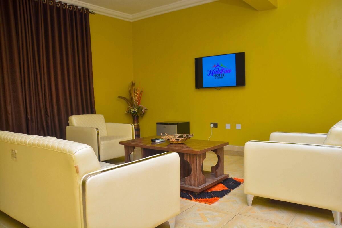A room with yellow walls, a television mounted on the wall, a wooden table, and two white armchairs.