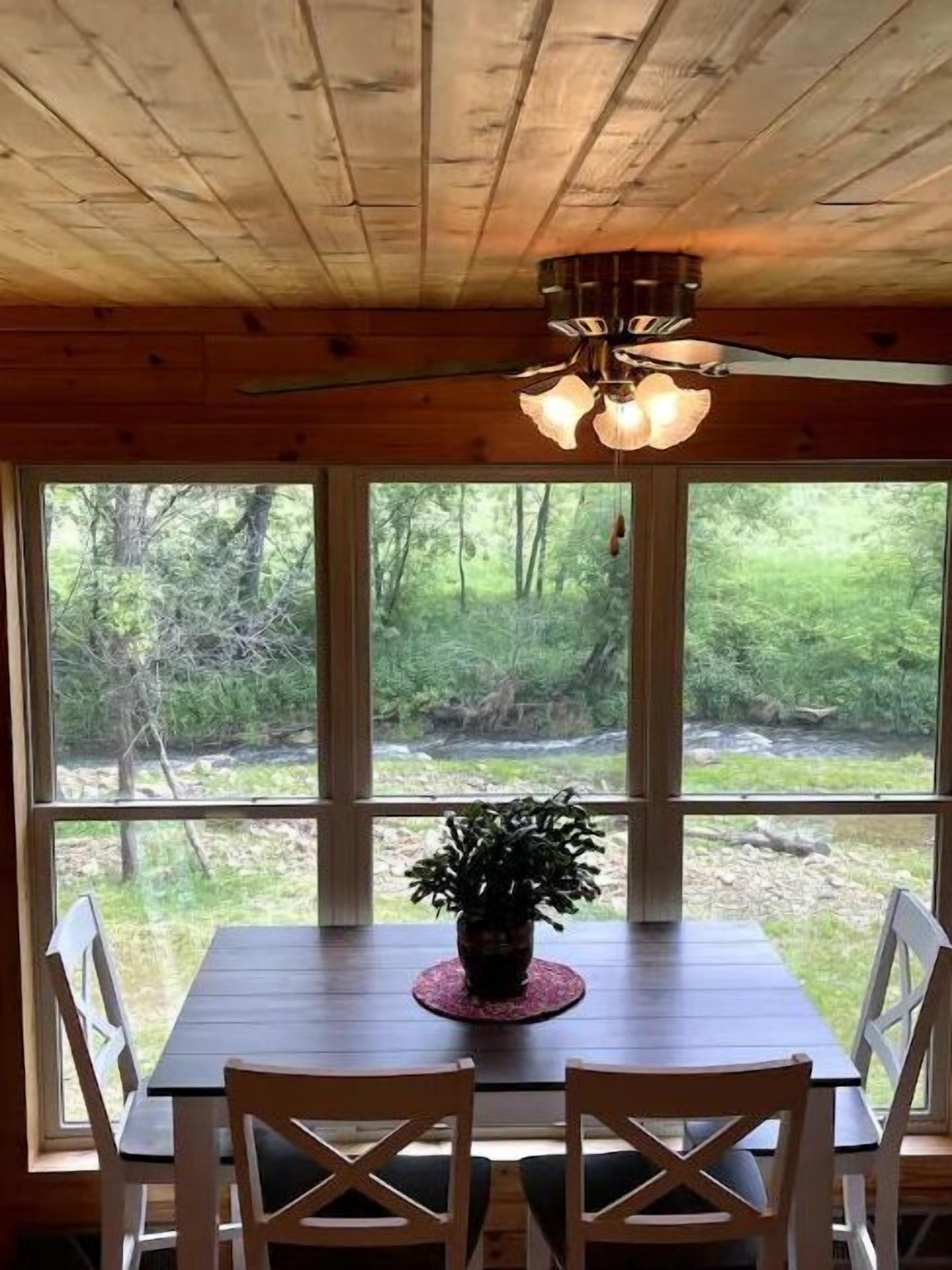 A wooden dining table with four chairs, a ceiling fan, and a view of a forest through large windows.
