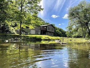 Surrounded by lush greenery, this riverside house offers a tranquil environment with a prominent entrance and balcony overlooking the calm river.