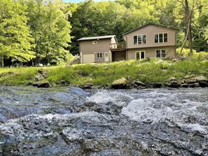 A two-story house with a garage, surrounded by lush greenery and a flowing stream. - Timber Coulee Creek in backyard, king size bed, grill on deck, large garage. (Westby)