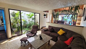 A living room with a brown sofa, a coffee table, and a chair. There is a sliding glass door leading to a balcony with plants.