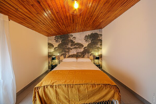 A bedroom with a wooden ceiling, a bed with a gold bedspread, two bedside lamps, and a mural of trees on the wall.