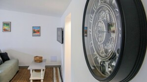 A room with a large wall clock, a small table with a basket, a sofa, and framed pictures on the wall. - Little House - GARDEN - GRAULHET (Graulhet)