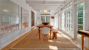 A spacious room with a wooden dining table, white chairs, and a large window providing a view of the outdoors. - Designer East Hampton Farmhouse - Bike to Amagansett, EH & Ocean Beaches, Pool (East Hampton)