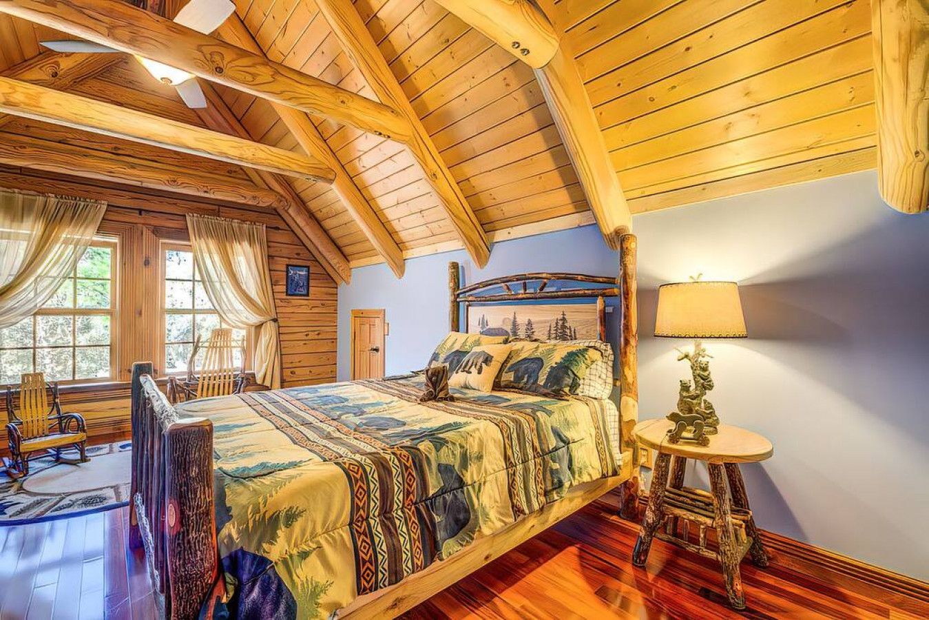 A cozy bedroom with a wooden ceiling, a large bed with a patterned bedspread, a bedside table with a lamp, and a window with curtains.