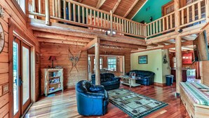 A spacious log cabin interior with a high ceiling, a balcony, and a cozy living area with leather sofas and a fireplace.