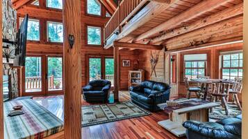 A spacious log cabin interior with a loft, wooden floors, a fireplace, and large windows.