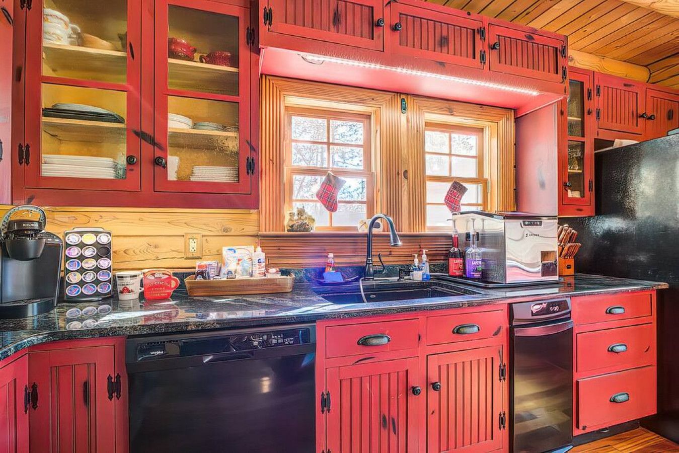 A kitchen with red cabinets, a black dishwasher, and a window with plaid curtains.