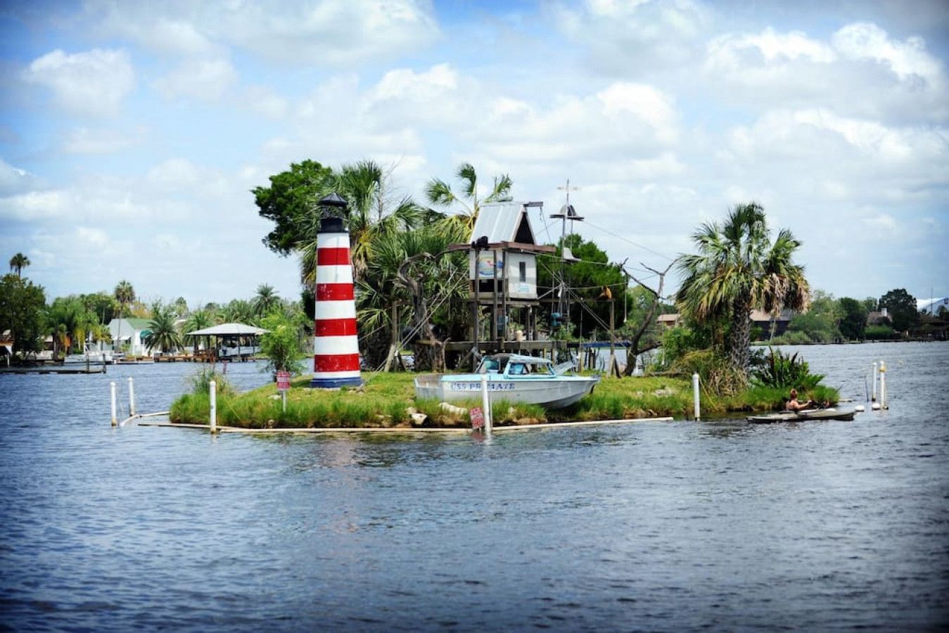 Una casa sull'acqua con un faro rosso e bianco a righe su una piccola isola, circondata da acqua e palme.