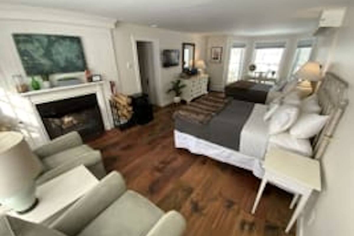 A modern bedroom with a large bed, a fireplace, a seating area with a sofa and armchairs, and a view of the outdoors.