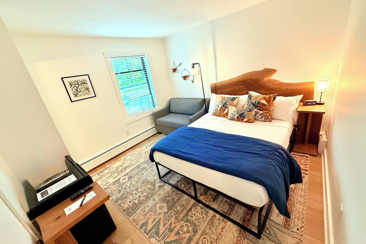A bedroom with a bed, a desk, a chair, a window with blinds, and a nightstand with a lamp.