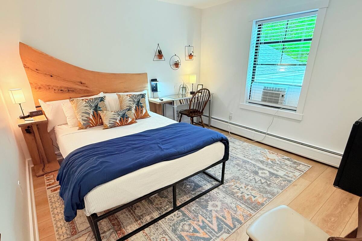 A modern bedroom with a large bed, a desk, and a chair. There is a window with blinds and a view of greenery outside.
