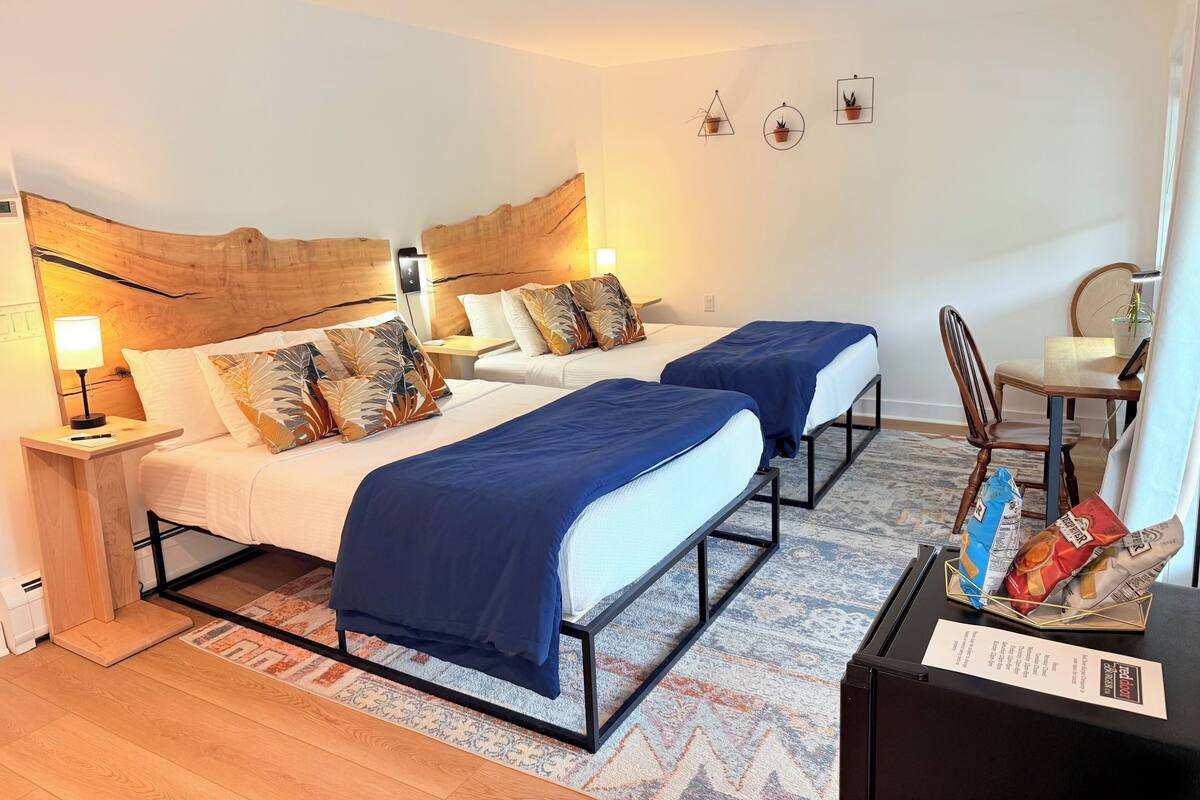 A hotel room with two beds, a wooden headboard, bedside lamps, a small table with a chair, and a wall-mounted plant.