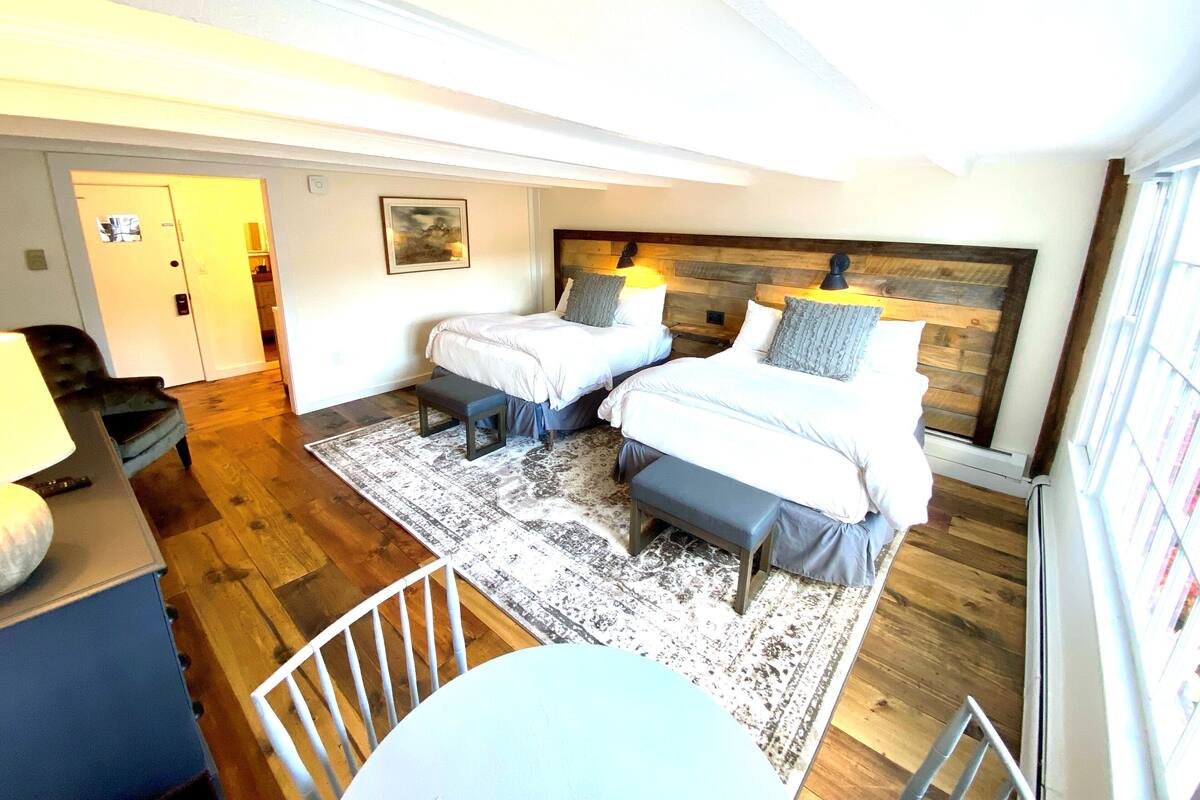 A bedroom with two beds, a desk, and a chair. There is a door leading to another room and a window with blinds.