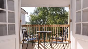 A wooden deck with a table and two chairs, a white door, and a view of trees and a building in the background.