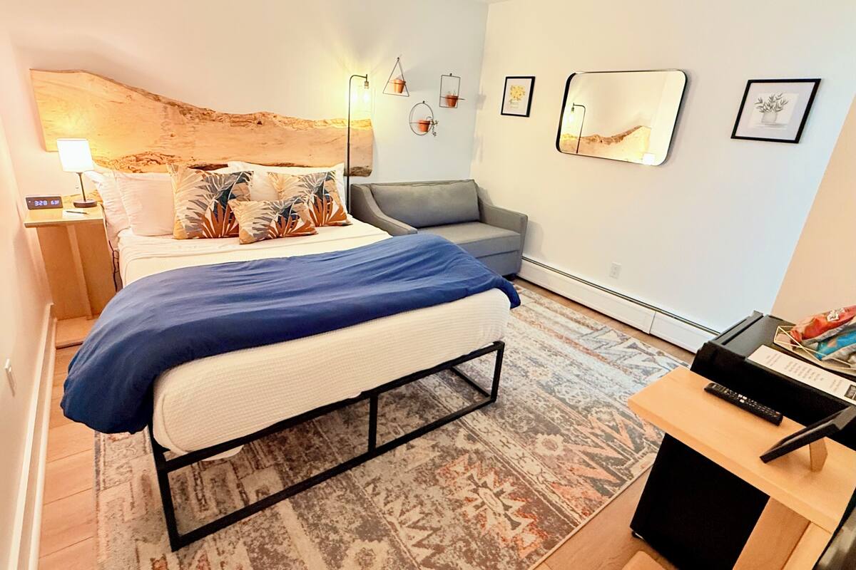 A modern bedroom with a bed, a grey sofa, a small table with a remote, and a wall-mounted mirror.