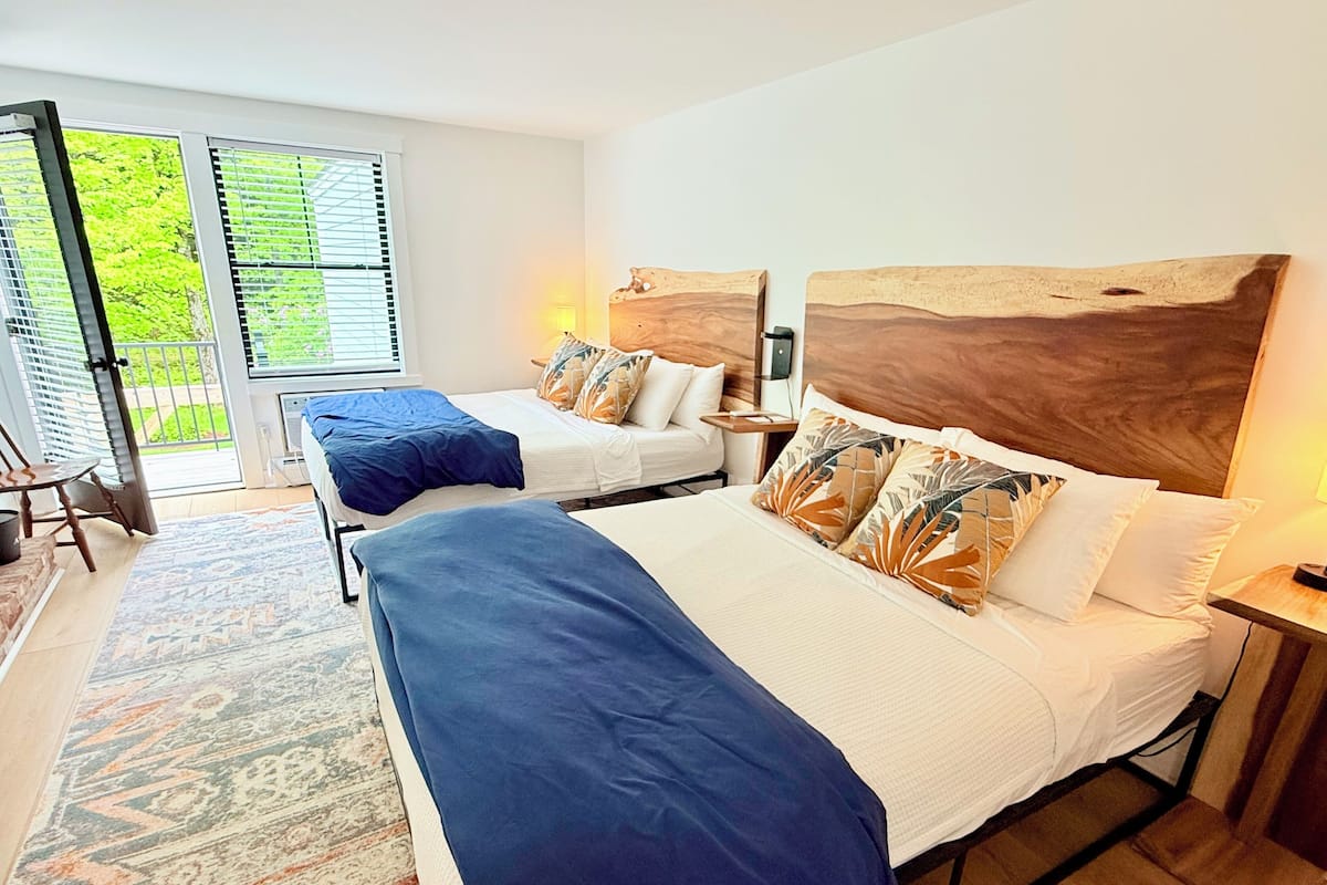 A hotel room with two beds, a large window with blinds, a patterned rug, and a wooden headboard.