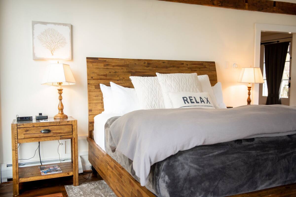 A bedroom with a wooden headboard, a bed with white bedding, a bedside table with a lamp, and a wall-mounted picture.