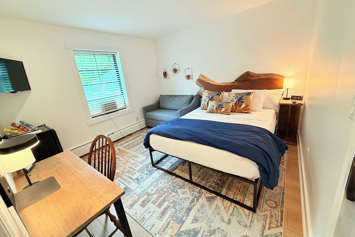A compact bedroom with a bed, bedside table, lamp, and a small seating area with a chair and sofa.