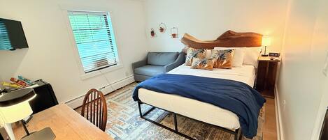 A compact bedroom with a bed, bedside table, lamp, and a small seating area with a chair and sofa.