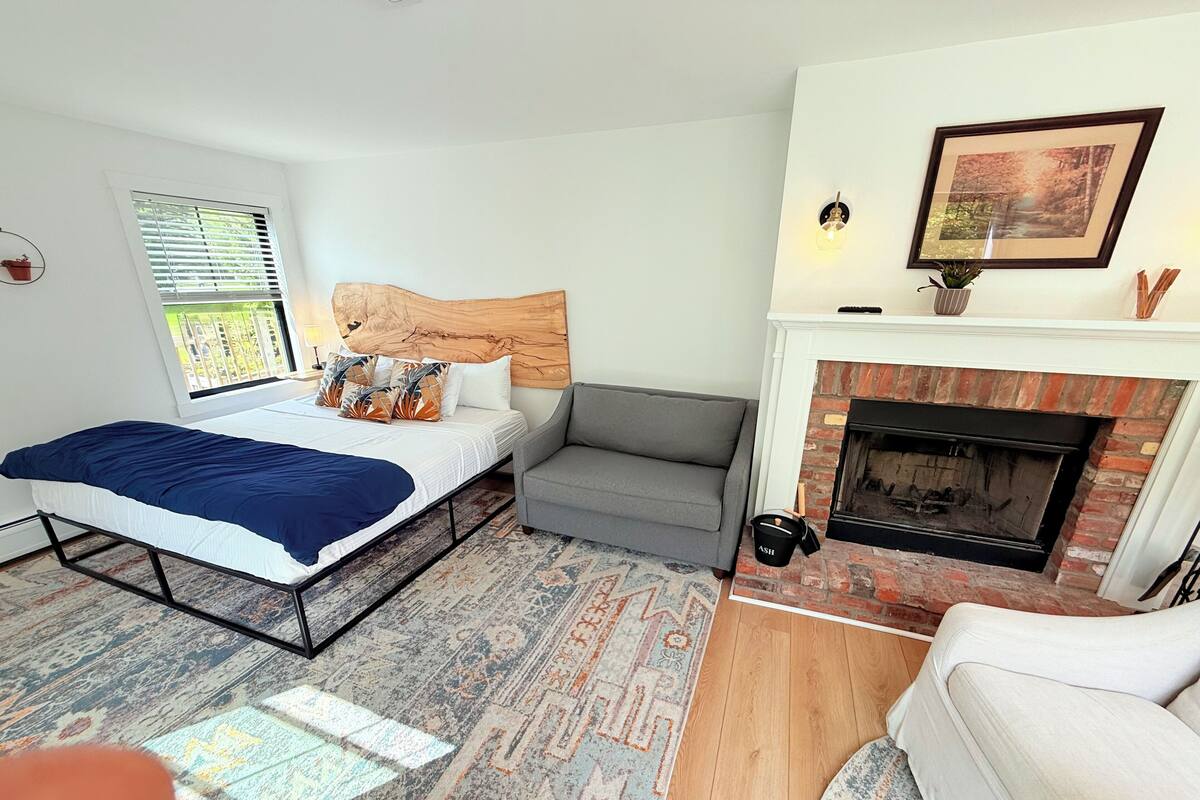 A bedroom with a bed, a grey sofa, a fireplace, and a patterned rug.