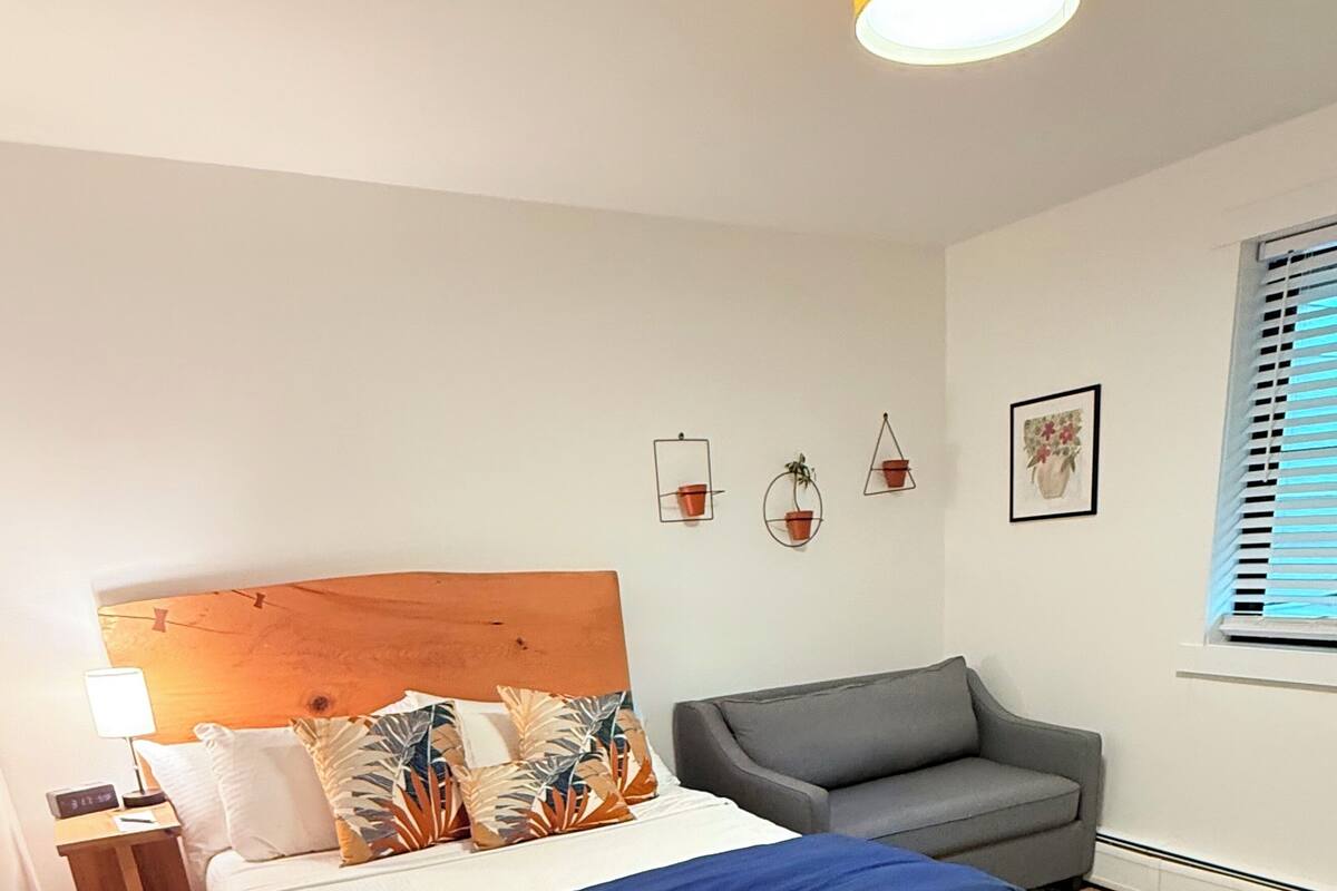 A modern bedroom with a wooden headboard, a bed with a blue blanket, a grey sofa, a patterned rug, and a small bedside table with a lamp.