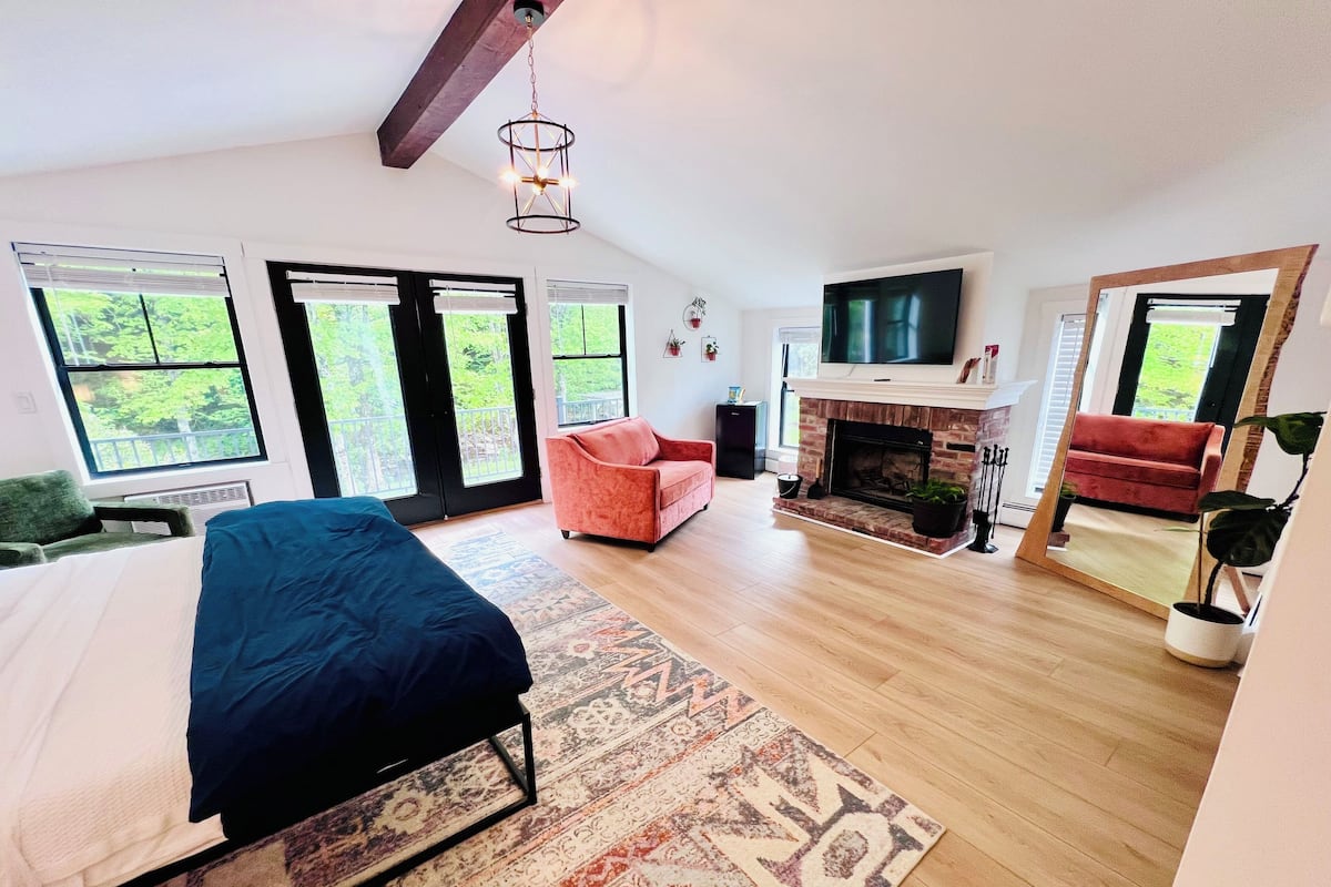 A spacious bedroom with a bed, a fireplace, a TV, and large windows with a view of greenery.