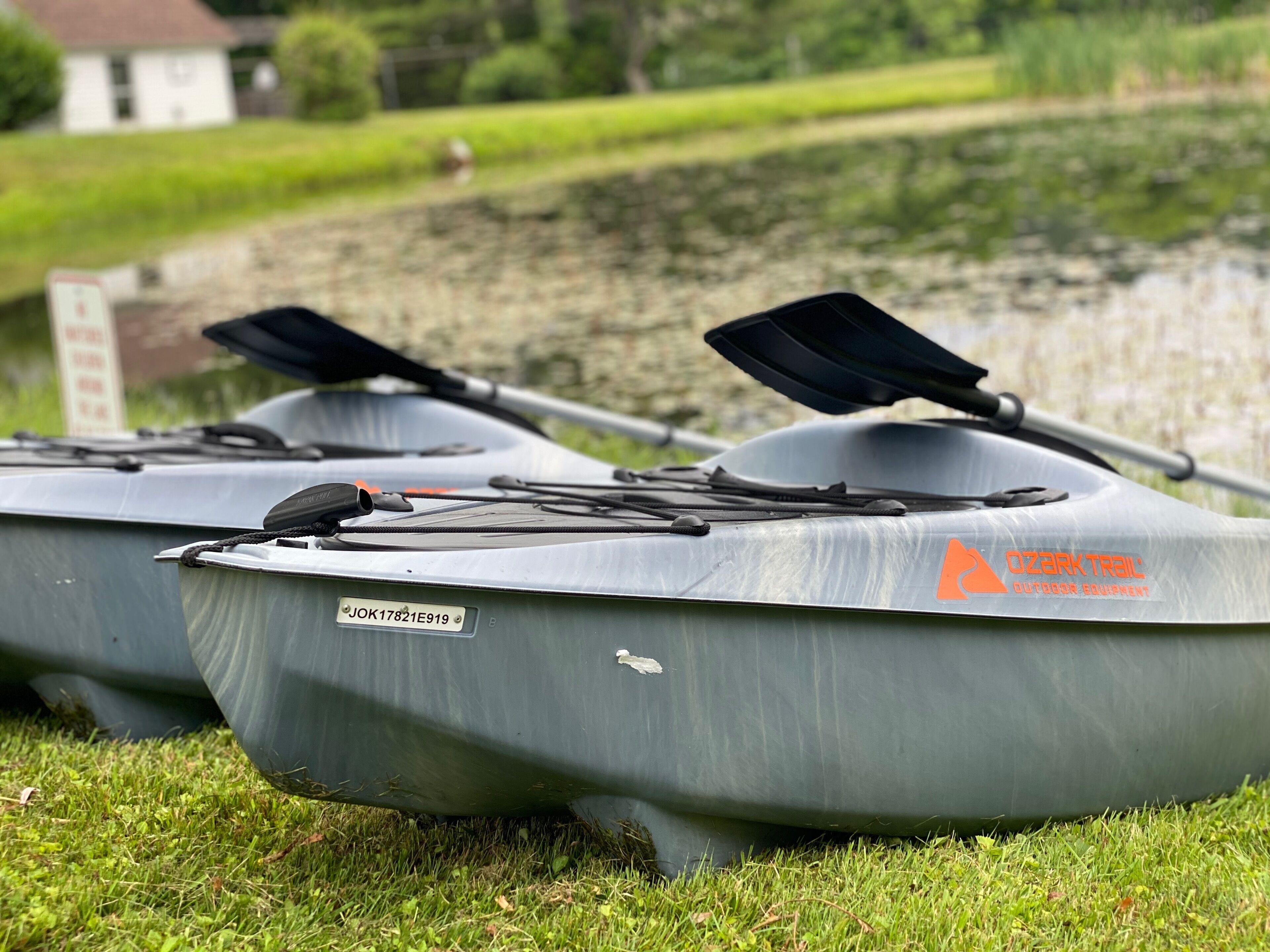 Two gray kayaks with 'OZARKTRAIL OUTDOOR EQUIPMENT' branding, parked on grass near a body of water.