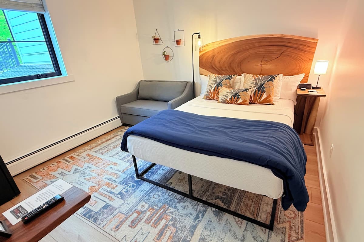 A bedroom with a bed, a grey sofa, a wooden headboard, a nightstand with a lamp, and a window with blinds.