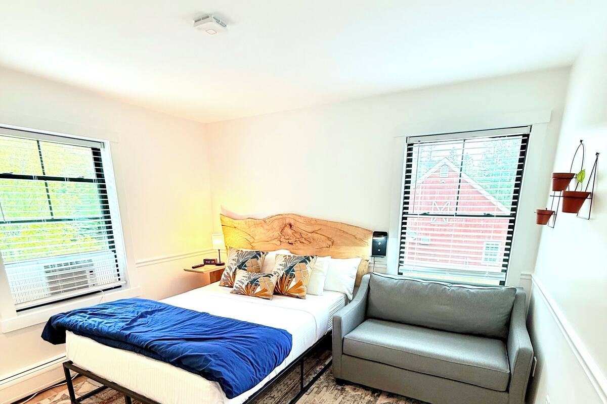 A bedroom with a bed, a gray sofa, a patterned rug, and large windows with blinds.