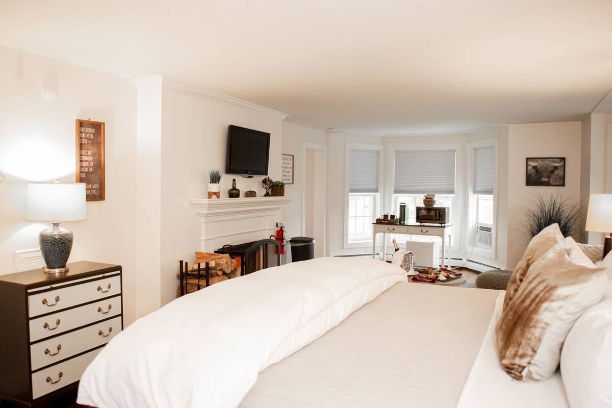 A bedroom with a large bed, a desk, a chair, a fireplace, and a television.