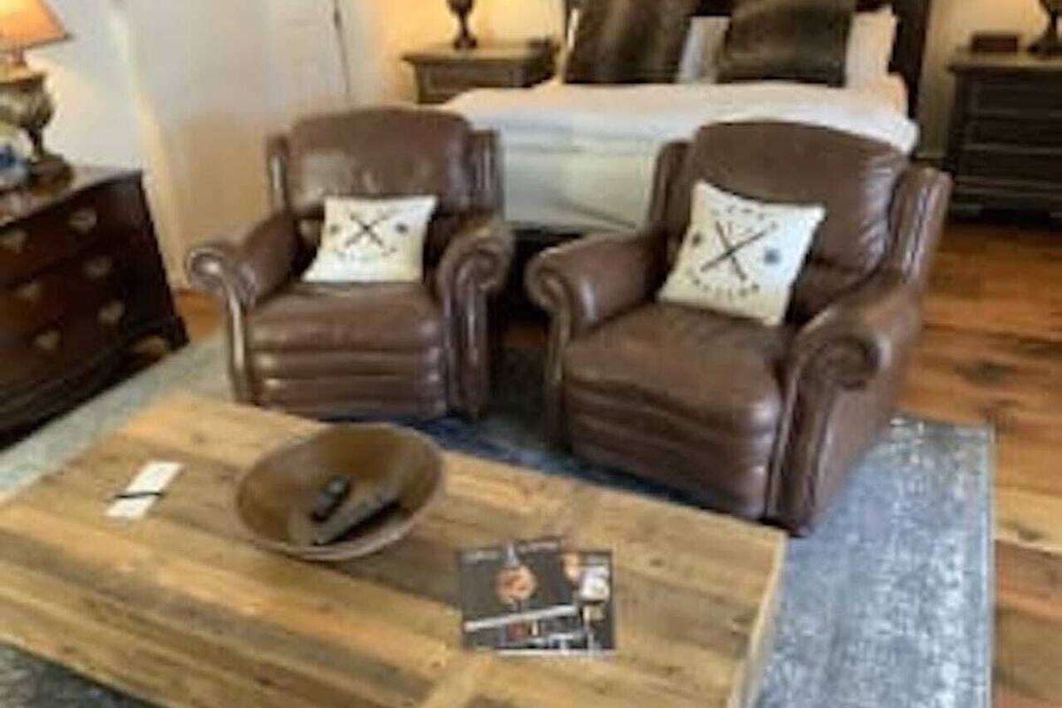 A living room with two brown leather armchairs, a wooden coffee table, and a bed with white bedding in the background.