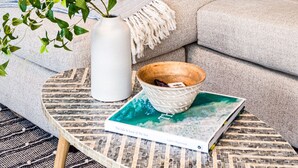 A living room with a beige sofa, blue and white cushions, a white vase, a wooden bowl, and a book on a round coffee table. - NEW! Drift Inn-Lakefront Home w/ Hot Tub, Outdoor Bar, Golf! (Lake Worth)