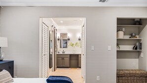 A bedroom with a bed, a nightstand, and a bedside table. There is a doorway leading to a bathroom with a toilet, sink, and mirror. The bathroom has a shower with a glass door. There is a small wicker basket on the floor. - NEW! Drift Inn-Lakefront Home w/ Hot Tub, Outdoor Bar, Golf! (Lake Worth)