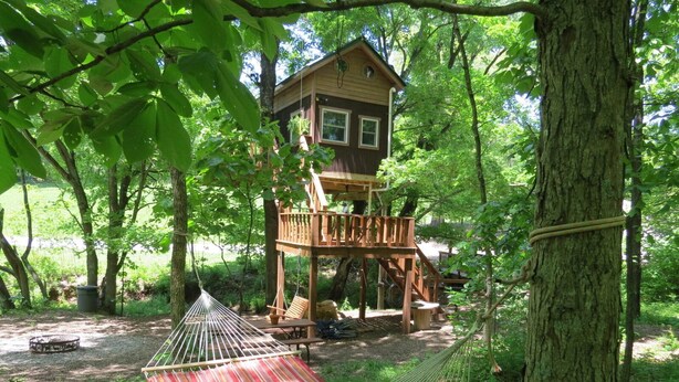 Una casa en el árbol con terraza de madera, una hamaca y una mesa de picnic.