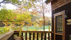 Una terraza de madera con una mesa y sillas, una puerta con un letrero de 'Welcome', y vistas a árboles otoñales y un campo verde.